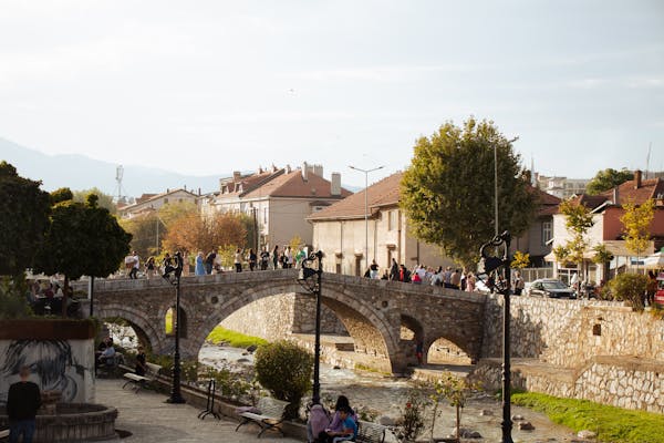 Scenic view of a historic stone bridge over a river in Prizren, Kosovo, with people walking and a charming urban backdrop.